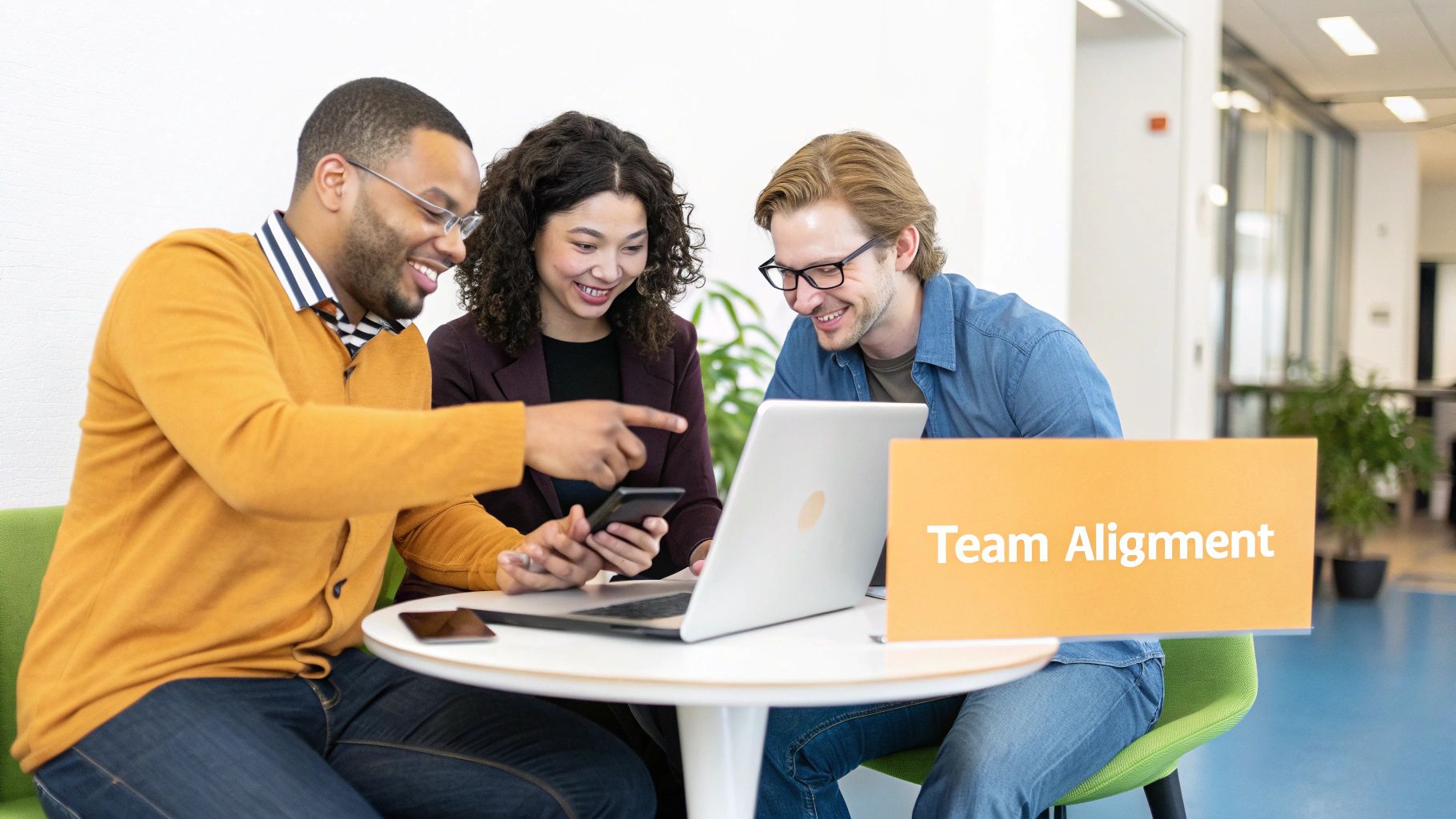 Three diverse colleagues smiling and collaborating around a laptop and phone, with a 'Team Alignment' sign.
