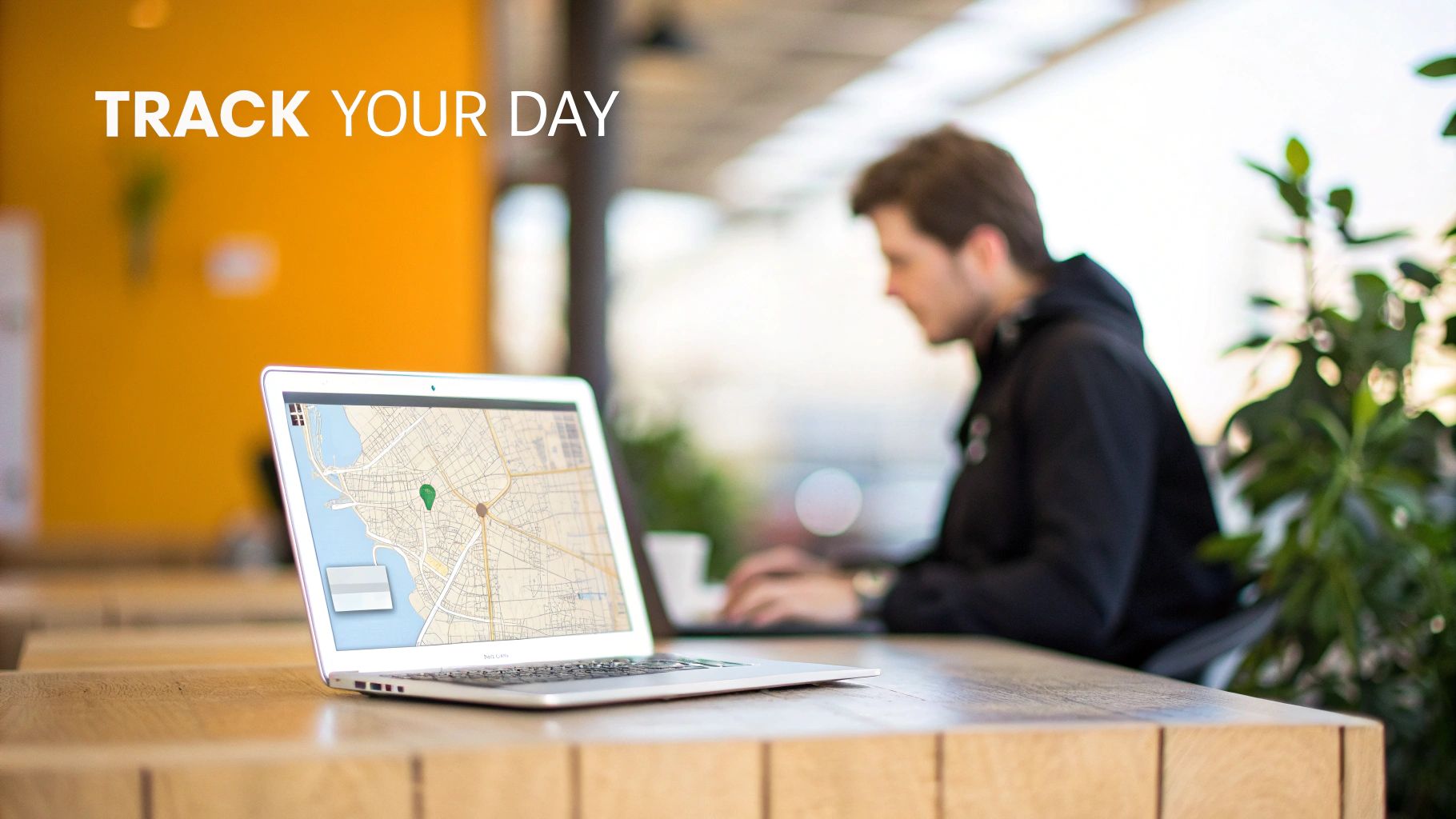 A laptop on a wooden table displays a map with a green location pin, with text 'TRACK YOUR DAY'.
